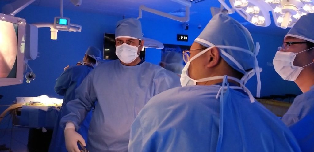 A team of surgeons in light blue surgical gowns and masks are performing a procedure in an operating room with blue-tinted lighting. A large monitor showing a live feed of the surgery is visible in the background.