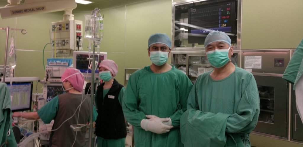 Two surgeons in green gowns, caps, and masks stand and smile for a photo in an operating room, with other medical staff and equipment visible in the background.