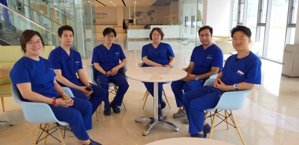 A group of six medical professionals, all wearing blue scrubs, sits around a small white table in a bright, modern-looking hospital lobby or waiting area.