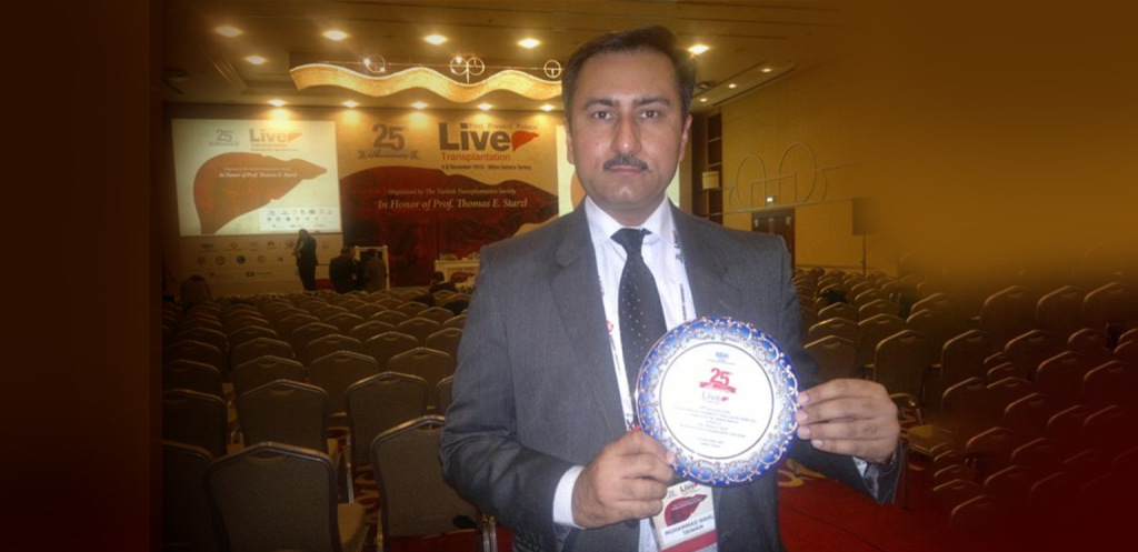 A doctor in a suit and tie holds a commemorative plate or award at a medical symposium. The background shows a stage with a banner for "Liver Transplantation," and rows of empty seats in an auditorium.