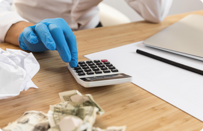 A person wearing a blue surgical glove uses a calculator on a wooden desk. Crumbled paper and crumpled dollar bills are on the desk, suggesting a focus on financial costs or medical billing.