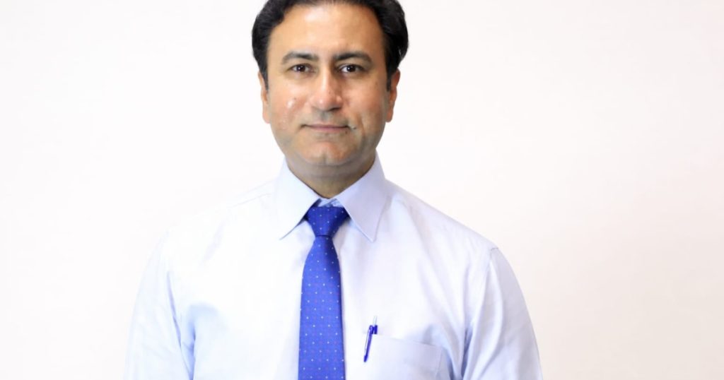 best cancer hospital in Lahore 2026 - A professional headshot of a middle-aged man with dark hair, wearing a light blue dress shirt and a blue patterned necktie against a plain white background.